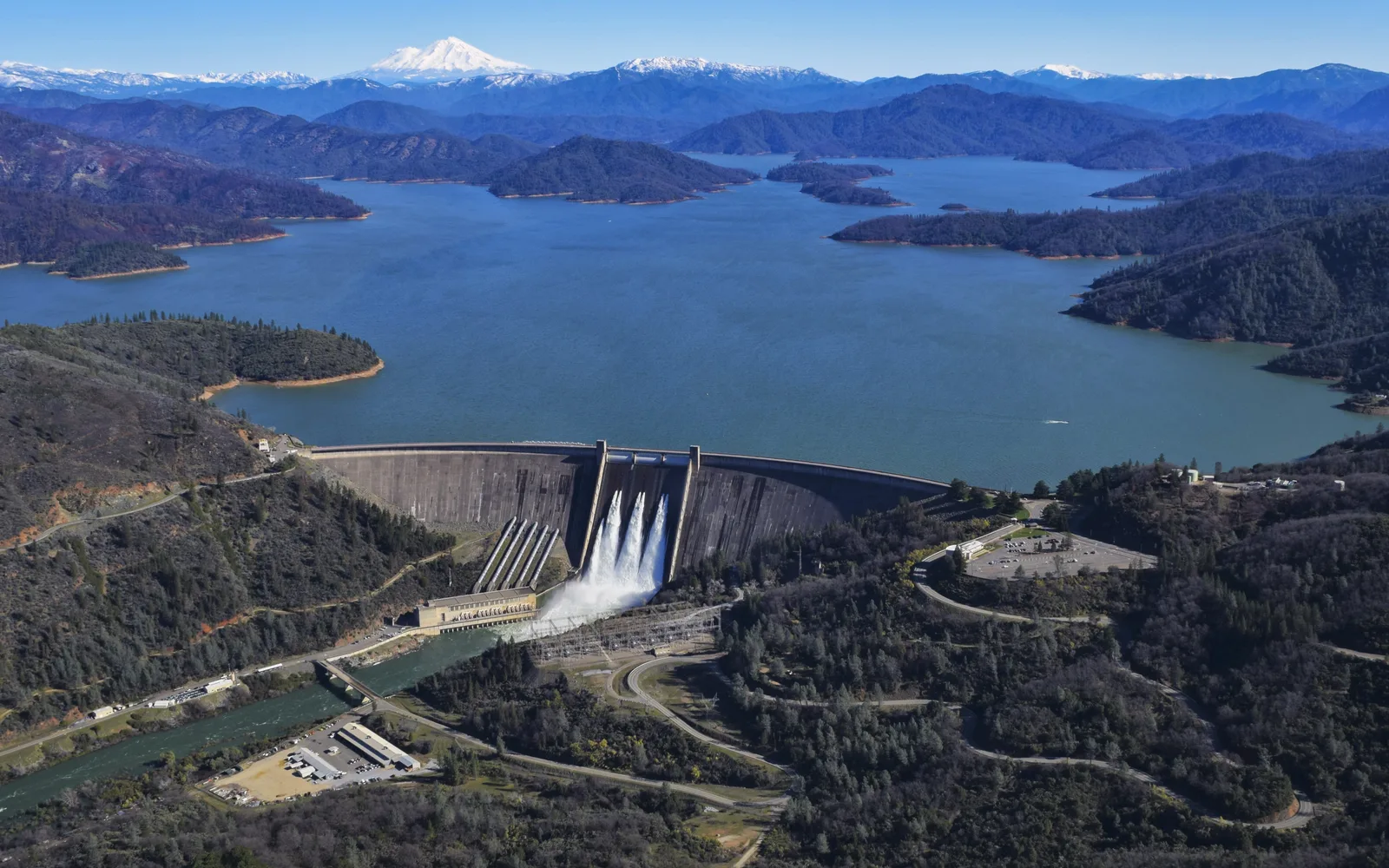 Aerial view of Shasta Dam, the concrete dam structure rising from the lake surrounded by green hills and blue water